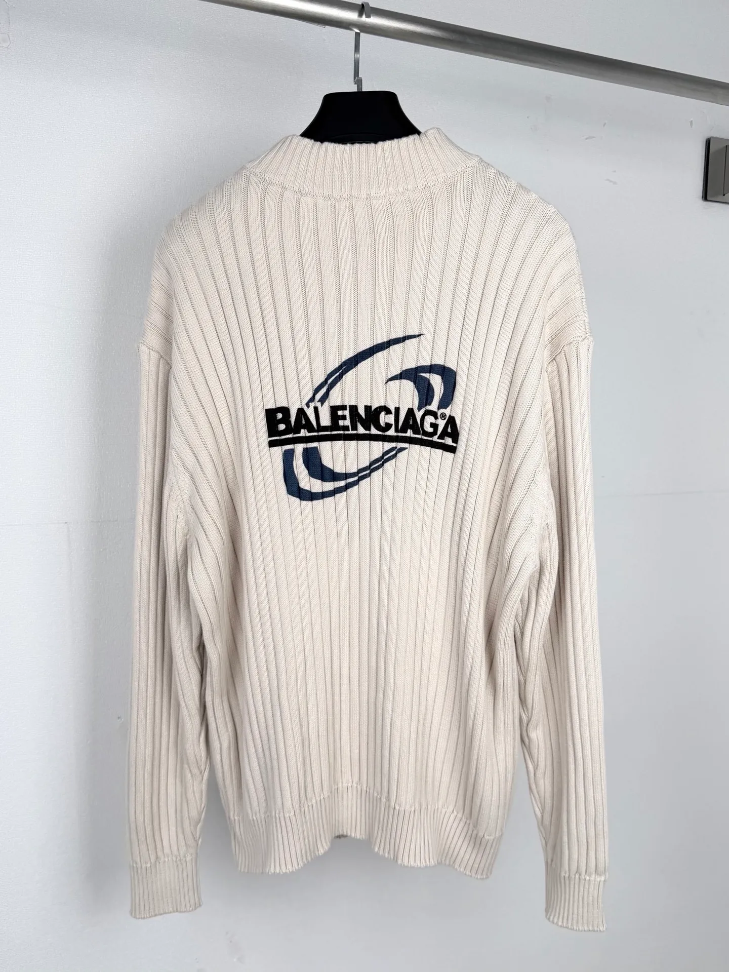 BALENCIAGA/Balenciaga Cola Wave Half-Zip Pit Knitted Sweater Ready-made garments are made of longitudinal pit texture knitted gray fabric. The weaving stage has high standards for yarn tension and machine yarn feeding speed. The width of the vertical grooves and the concave and convex texture of the entire body of the garment must be highly uniform. The production process is prone to defects such as texture deviation and density gaps, and the gray fabric screening loss is large. After the gray fabric is off the machine, it is pre-shrunk and heat-set to release the potential shrinkage of the fabric in advance and prevent the subsequent distortion and size deformation of the finished garments in the washing water. The body of the garment is equipped with a half-zip stand-up collar structure. The collar ribs and body pit stripes require vertical alignment, and the splicing process requires precise alignment of the texture direction. The zipper welt uses a blind-stitching process to ensure smooth opening and closing of the zipper and avoid local vertical grain stretch deformation caused by sewing pull. Sewing workers need to continuously control the fabric feeding tension to balance the elasticity of the fabric and the stability of the threading. Drop-shoulder silhouette pieces are cut strictly along the pitted texture to prevent diagonal cuts from damaging the fabric texture. The printing of the wave monogram on the back is the core technical difficulty of this model. The surface of the concave and convex pit fabric is undulating and cannot be printed directly like flat fabrics. During production, the tension of the printing area is first fixed and leveled, and the layered alignment offset printing process is used to complete the printing of wave graphics and letter logos in sequence. Multi-layer pattern overprinting requires extremely high precision, and it is easy to produce ghosting and misalignment defects. The ink ratio has been adjusted to take into account the fullness of the pattern and ensure that the printing is not prone to cracking and falling off when the fabric is stretched. The cuffs and hem ribs are woven from pitted fabrics of the same system, and the elastic density matches the body parameters to avoid problems with elasticity imbalance and wavy edges after wearing. After the garments are sewn, detailed inspection is carried out to screen for sewing defects and printing defects. Finally, the low-temperature wet finishing process is used to uniformly correct the pattern and texture flatness. From knitting and weaving, precision cutting, structural sewing, multi-layer overprinting to garment styling, the process chain is complicated. Failure to control any process standard will directly affect the overall look and feel of the garment. Size:XS S M L